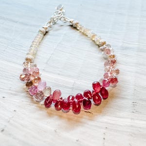 May include: A delicate bracelet featuring a gradient of pink, red, and white gemstone beads. The bracelet is crafted with silver clasps and showcases a range of bead sizes and shapes.