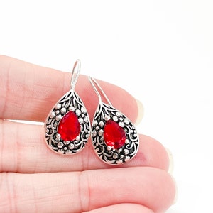 May include: A pair of silver teardrop earrings featuring a vibrant red gemstone. The earrings have an intricate floral pattern surrounding the stone and a hook fastening. The earrings are shown against a white backdrop.