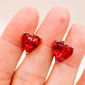 Peut inclure: Une paire de boucles d'oreilles à clous en forme de cœur. Chaque boucle d'oreille présente une pierre précieuse rouge vif à facettes sertie dans un métal argenté. Les boucles d'oreilles sont présentées sur un fond clair.