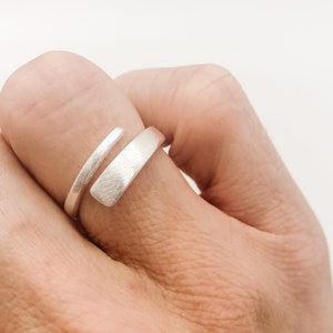 May include: A silver ring with a contemporary, minimalist design. The ring has a wrap-around style with a textured, rectangular band and a smooth, thin band. The ring is worn on a finger.