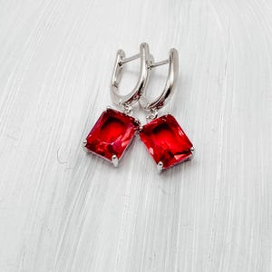 May include: A pair of silver-toned earrings with a lever back closure. Each earring features a large, rectangular, vibrant red gemstone, set in a silver bezel. The gemstones have a faceted cut, reflecting light.