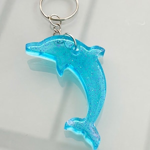 Puede incluir: Un llavero de delfín azul con purpurina incrustada en el plástico. El delfín está saltando fuera del agua y tiene un llavero plateado.