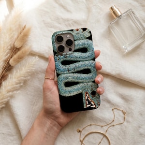 Op de afbeelding: Een zwarte smartphonehoes met een textuurrijk, lichtblauw, kronkelend slangontwerp met een gedetailleerde kop en witte tanden. De hoes heeft een uitsparing voor de cameralens aan de linkerbovenkant van het beschermende accessoire.