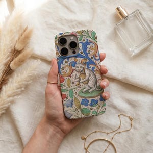 May include: A smartphone case featuring a detailed illustration of a monkey playing a musical instrument, surrounded by intricate blue, green, and orange floral and vine patterns on a cream-colored background with a camera cutout at the top.