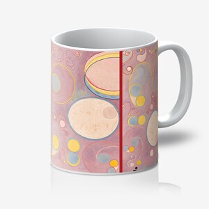 Puede incluir: Taza de café de cerámica blanca con un diseño abstracto colorido. El diseño incluye varias formas en tonos de rosa, amarillo, azul y blanco, con una franja roja vertical. La taza tiene un asa e interior blancos.