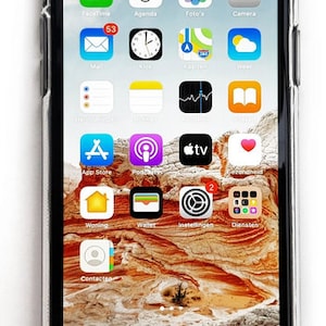 May include: A close-up of an iPhone screen displaying various apps, including FaceTime, Agenda, Photos, Camera, Mail, Clock, Maps, Weather, App Store, Podcasts, Apple TV, and more. The phone is in a clear case and the background is a blurry image of a desert landscape.