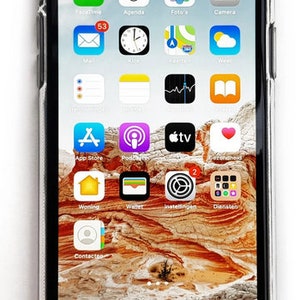 Puede incluir: Primer plano de la pantalla de un iPhone que muestra varias aplicaciones, incluyendo FaceTime, Agenda, Fotos, C&aacute;mara, Correo, Reloj, Mapas, Clima, App Store, Podcasts, Apple TV, y m&aacute;s. El tel&eacute;fono est&aacute; en una funda transparente y el fondo es una imagen borrosa de un paisaje des&eacute;rtico.