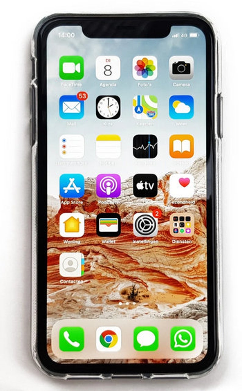 Puede incluir: Primer plano de la pantalla de un iPhone que muestra varias aplicaciones, incluyendo FaceTime, Agenda, Fotos, C&aacute;mara, Correo, Reloj, Mapas, Clima, App Store, Podcasts, Apple TV, y m&aacute;s. El tel&eacute;fono est&aacute; en una funda transparente y el fondo es una imagen borrosa de un paisaje des&eacute;rtico.