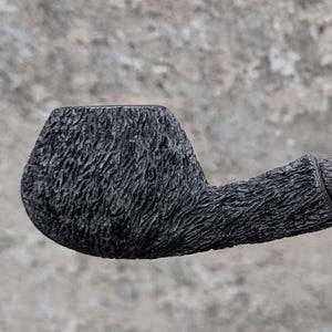 May include: A black smoking pipe with a textured, rough surface. The pipe has a curved stem and a metal tip. The bowl and stem are connected by a smooth, cylindrical section. The pipe is designed for smoking tobacco.