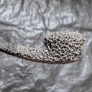 Könnte beinhalten: Eine graue Pfeife mit einem strukturierten, porösen Kopf und Stiel. Die Pfeife hat einen dunklen, schlanken Stiel und einen Kopf mit einer rauen, zerfurchten Oberfläche. Die Pfeife ist zum Rauchen bestimmt.