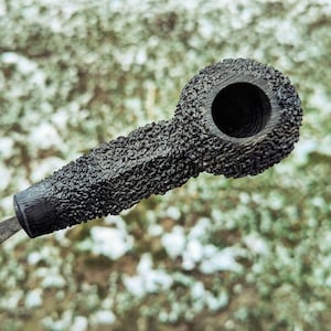 Op de afbeelding: Close-up van een zwarte tabakspijp. De pijp heeft een ruwe, schorsachtige textuur op de kop en steel, in contrast met een glad, donker mondstuk. De kop heeft een grote, cirkelvormige opening.