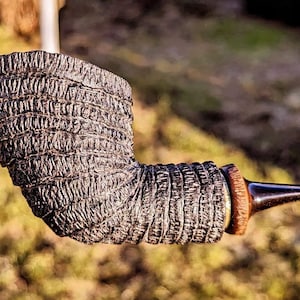 May include: A dark brown, textured tobacco pipe with a curved stem and a smooth, dark mouthpiece. The pipe's bowl has a layered, ridged design. The background is blurred, suggesting an outdoor setting.
