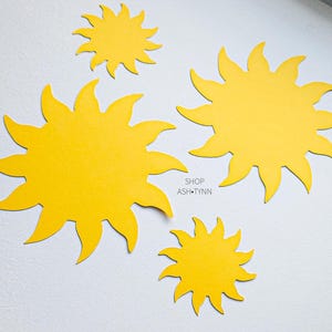 May include: Four yellow sun cutouts of varying sizes are arranged on a white surface. The suns have pointed rays and a textured appearance. The text "SHOP ASH.TYNN" is visible.