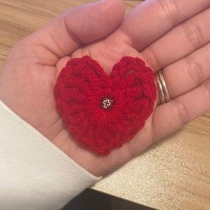 May include: A handmade red crocheted heart with a small silver cross charm in the center. The heart is held in a person's hand, showcasing the texture of the yarn and the delicate charm. The heart is a symbol of love and faith.