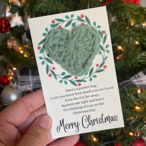 May include: A greeting card with a green crocheted heart, surrounded by a wreath of green leaves and red berries. The card includes the text "Here's a pocket hug...Merry Christmas."