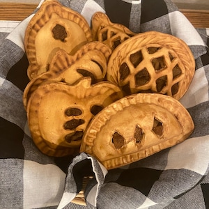 May include: A basket filled with ceramic pies in various shapes, including a lattice-top pie and several with cut-out designs. The pies are a warm tan color with darker brown accents, resting on a black and white checkered cloth.