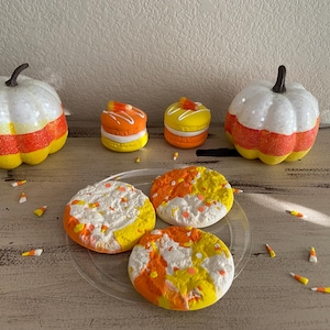 May include: Four Halloween-themed cookies decorated with orange, yellow, and white frosting. The cookies are on a clear plate and surrounded by candy corn. Two white pumpkins with orange and yellow stripes are in the background.