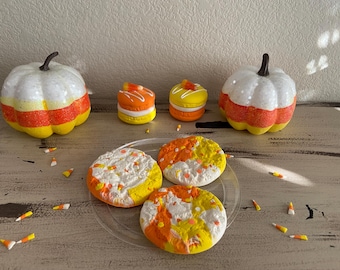 Puede incluir: Cuatro galletas con temática de Halloween decoradas con glaseado naranja, amarillo y blanco. Las galletas están en un plato transparente y rodeadas de maíz dulce. Dos calabazas blancas con rayas naranjas y amarillas están en el fondo.