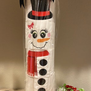 May include: A snowman-shaped gift wrap made from toilet paper rolls. The snowman has a black top hat, red scarf, and black buttons. The snowman is wrapped in clear plastic and tied with a black ribbon.