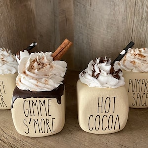 May include: Four ceramic mugs with whipped cream and toppings, decorated with fall-themed text. The mugs are labeled "Coffee Bar", "Gimme S'more", "Hot Cocoa", and "Pumpkin Spice".