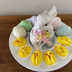 May include: A white ceramic bunny figurine holding a bouquet of pink and yellow flowers sits in the center of a white egg holder. The egg holder is filled with six deviled eggs, each topped with yellow yolk and paprika. The bunny is surrounded by pastel-colored Easter eggs.