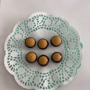 May include: Six chocolate-covered peanut butter Buckeyes arranged on a white and mint green paper doily. The candies have a brown chocolate coating with a tan peanut butter center. The doily has a floral lace design.