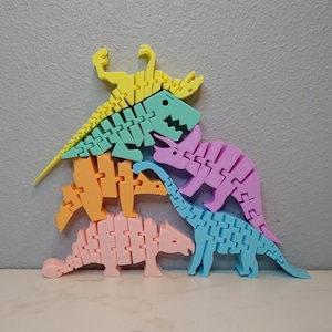 May include: A set of five colorful dinosaur skeletons made of plastic. The dinosaurs are stacked on top of each other, with the largest dinosaur at the bottom and the smallest dinosaur at the top. The dinosaurs are all different colors, including pink, blue, green, orange, and yellow.