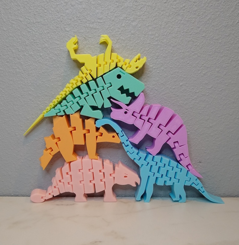May include: A set of five colorful dinosaur skeletons made of plastic. The dinosaurs are stacked on top of each other, with the largest dinosaur at the bottom and the smallest dinosaur at the top. The dinosaurs are all different colors, including pink, blue, green, orange, and yellow.