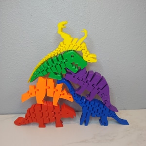 May include: A set of five colorful dinosaur toys made of plastic. The dinosaurs are stacked on top of each other, with the yellow dinosaur on top and the blue dinosaur on the bottom. The dinosaurs are all different colors and have different shapes.