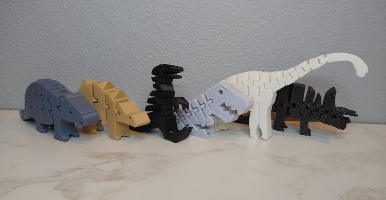 May include: Five articulated dinosaur toys made of plastic. The dinosaurs are in a row from left to right: a blue ankylosaurus, a tan ankylosaurus, a black tyrannosaurus rex, a white stegosaurus, and a black triceratops.