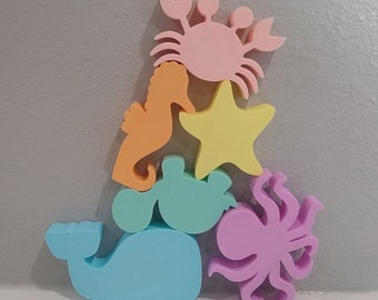 May include: A stack of colorful sea creature shaped foam toys. The bottom toy is a light blue whale, followed by a light green sea turtle, a yellow starfish, an orange seahorse, and a pink octopus.