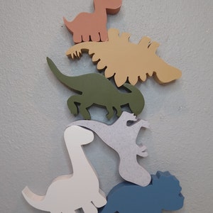 Puede incluir: Una pila de cinco figuritas de dinosaurio de madera en varios colores, incluyendo verde, azul, blanco, rosa y beige. Los dinosaurios están dispuestos en forma de pirámide, con el dinosaurio más pequeño en la parte superior y el dinosaurio más grande en la parte inferior.