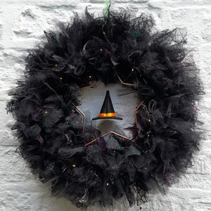 May include: A black decorative wreath constructed from layers of tulle and mesh fabric, featuring a copper-coloured star frame. A small black witch hat with an orange band hangs in the centre. The wreath is illuminated by small, warm-toned LED lights.