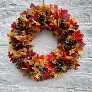 May include: A vibrant autumn wreath featuring a circular design. The wreath is adorned with colorful fall leaves in shades of red, orange, and yellow. Pine cones, chestnuts, and small red and white mushroom decorations are also incorporated, creating a festive seasonal display.