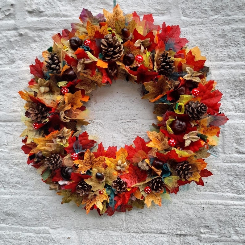 May include: A vibrant autumn wreath featuring a circular design. The wreath is adorned with colorful fall leaves in shades of red, orange, and yellow. Pine cones, chestnuts, and small red and white mushroom decorations are also incorporated, creating a festive seasonal display.