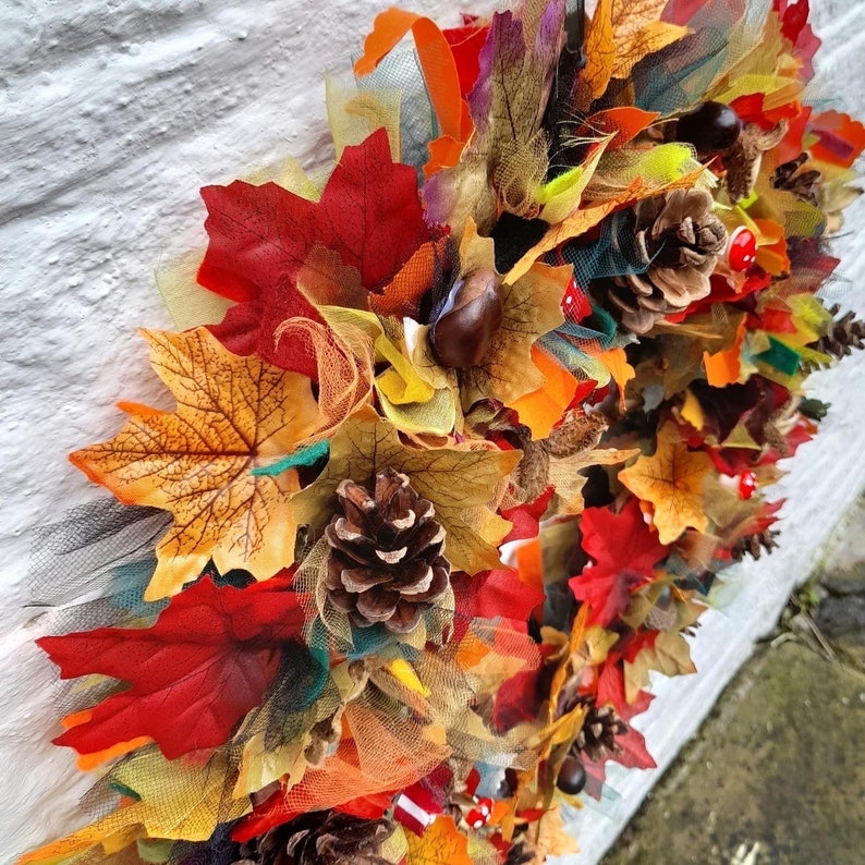 May include: A colorful autumn-themed garland featuring artificial leaves in shades of red, orange, and yellow. The garland also includes pine cones, chestnuts, and decorative tulle, creating a festive fall decoration.