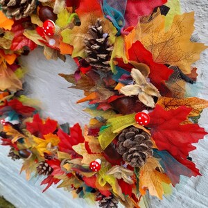 May include: A colorful autumn wreath featuring red, orange, and yellow fabric leaves, pinecones, and small red mushroom accents. The wreath is decorated with various textures and colors, creating a festive fall decoration.