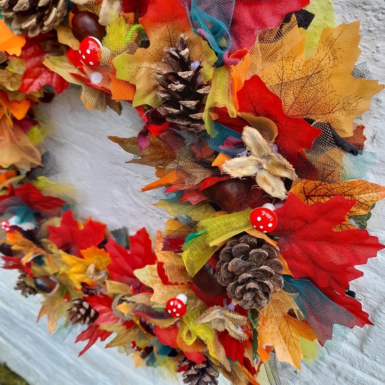 May include: A colorful autumn wreath featuring red, orange, and yellow fabric leaves, pinecones, and small red mushroom accents. The wreath is decorated with various textures and colors, creating a festive fall decoration.