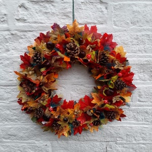 May include: A vibrant autumn wreath featuring a circular design. The wreath is adorned with colorful fabric leaves in shades of red, orange, and yellow, along with pine cones, chestnuts, and small red mushroom accents. It hangs against a white brick wall.