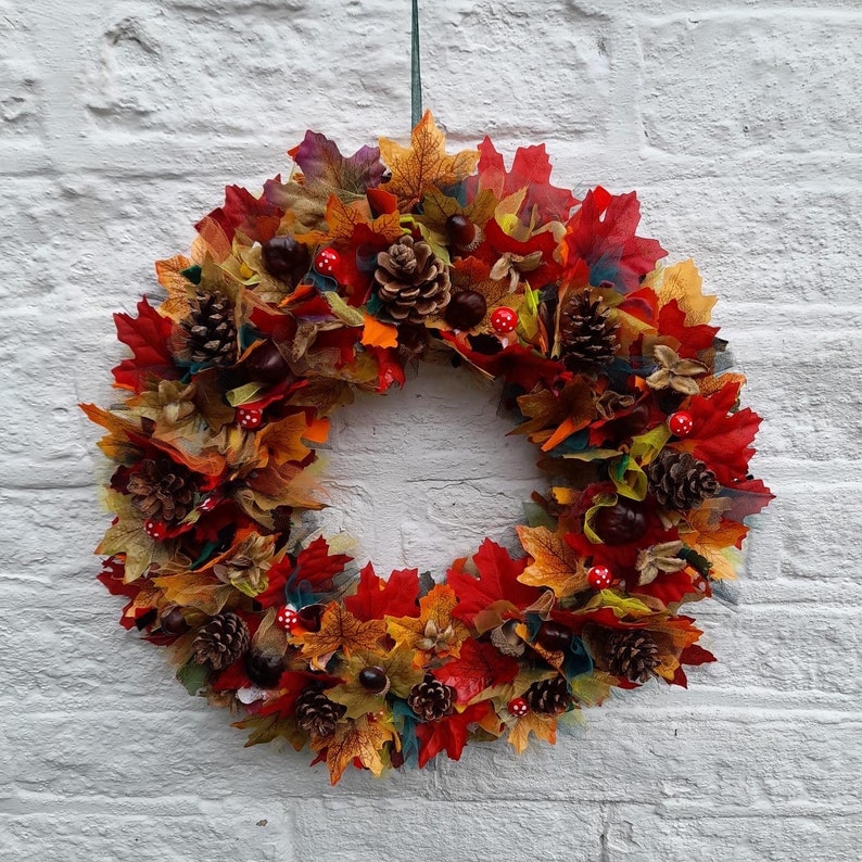 May include: A vibrant autumn wreath featuring a circular design. The wreath is adorned with colorful fabric leaves in shades of red, orange, and yellow, along with pine cones, chestnuts, and small red mushroom accents. It hangs against a white brick wall.