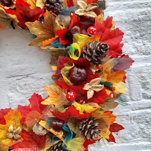 May include: A vibrant autumn wreath featuring colorful faux leaves in shades of red, orange, and yellow. The wreath is adorned with pinecones, chestnuts, and small red and white mushroom accents, creating a festive fall theme.