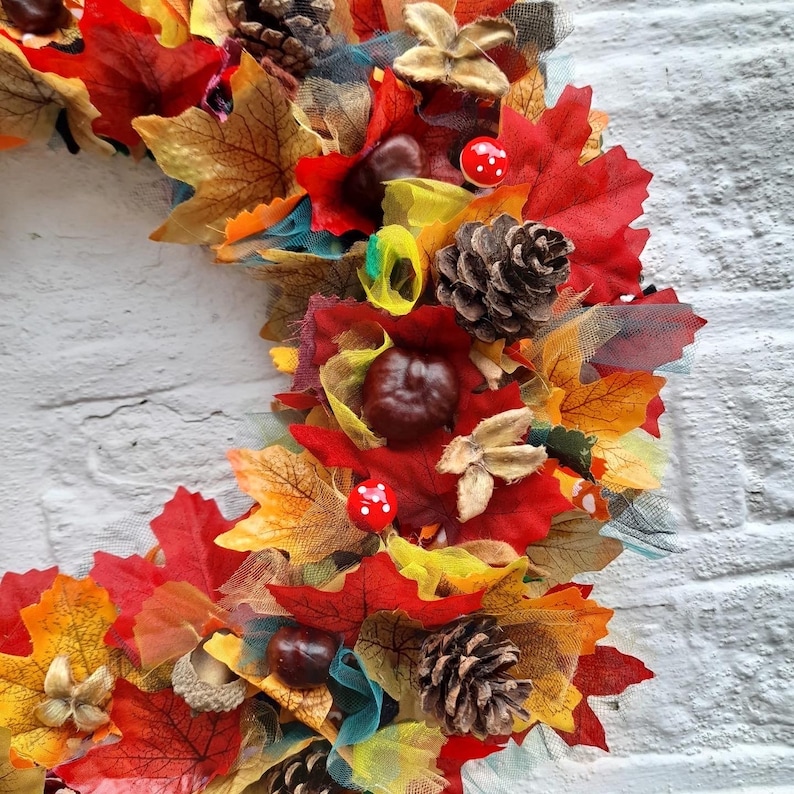 May include: A vibrant autumn wreath featuring colorful faux leaves in shades of red, orange, and yellow. The wreath is adorned with pinecones, chestnuts, and small red and white mushroom accents, creating a festive fall theme.