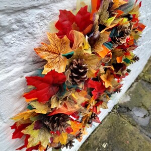 May include: A fall-themed decorative garland featuring vibrant red, orange, and yellow artificial leaves, pine cones, and accents of green and brown. The garland is arranged against a white textured wall, creating a seasonal display.