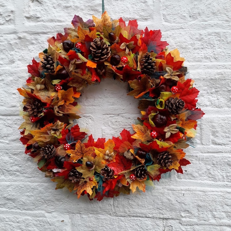 May include: A circular autumn wreath composed of colorful leaves in shades of red, orange, and yellow. The wreath is adorned with pine cones, small red and white mushrooms, and chestnuts, creating a festive fall decoration.