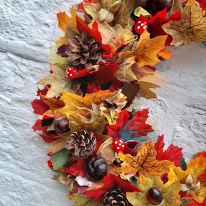 May include: A fall-themed wreath with a variety of autumn leaves in shades of red, orange, and yellow. The wreath also features pine cones, chestnuts, and small red mushrooms with white spots. The wreath is set against a white brick wall.