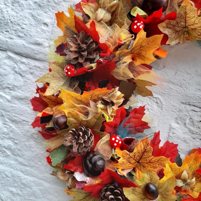 May include: A fall-themed wreath with a variety of autumn leaves in shades of red, orange, and yellow. The wreath also features pine cones, chestnuts, and small red mushrooms with white spots. The wreath is set against a white brick wall.