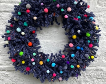 May include: A navy blue yarn wreath decorated with colorful pom-poms. The wreath is hanging on a white brick wall.