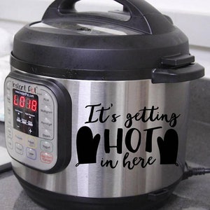 Puede incluir: Una olla a presión eléctrica Instant Pot de acero inoxidable con tapa y base negras. El frente presenta un panel de control digital y una calcomanía de vinilo negra que dice "It's getting HOT in here" flanqueada por dos iconos de guantes.
