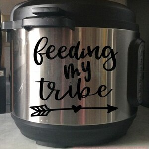 Peut inclure: Un autocuiseur électrique en acier inoxydable avec un autocollant en vinyle noir sur le devant. L'autocollant affiche le texte "feeding my tribe" dans une police cursive, placé au-dessus d'un graphique de flèche horizontale avec un petit cœur.