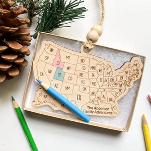 May include: Wooden United States map ornament with state outlines and a blue pencil. The ornament is in a gift box with the text "The Anderson Family Adventures". The states of Idaho and Utah are highlighted.